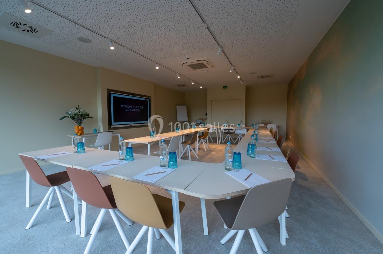 Salle de réunion lumineuse avec tables en U, chaises colorées, bouteilles d'eau et écran de présentation allumé.