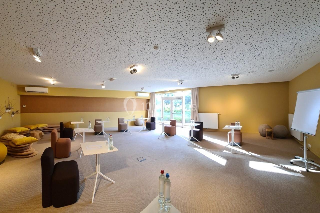 Salle de réunion lumineuse avec tables espacées, chaises, tableau blanc et bouteilles d'eau, vue sur l'extérieur.