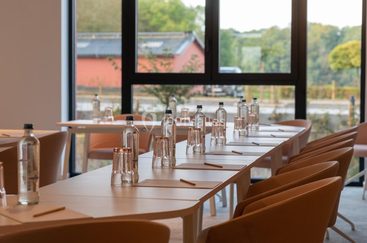Salle de réunion lumineuse avec des tables disposées en U, équipées de bouteilles d'eau, verres et crayons.
