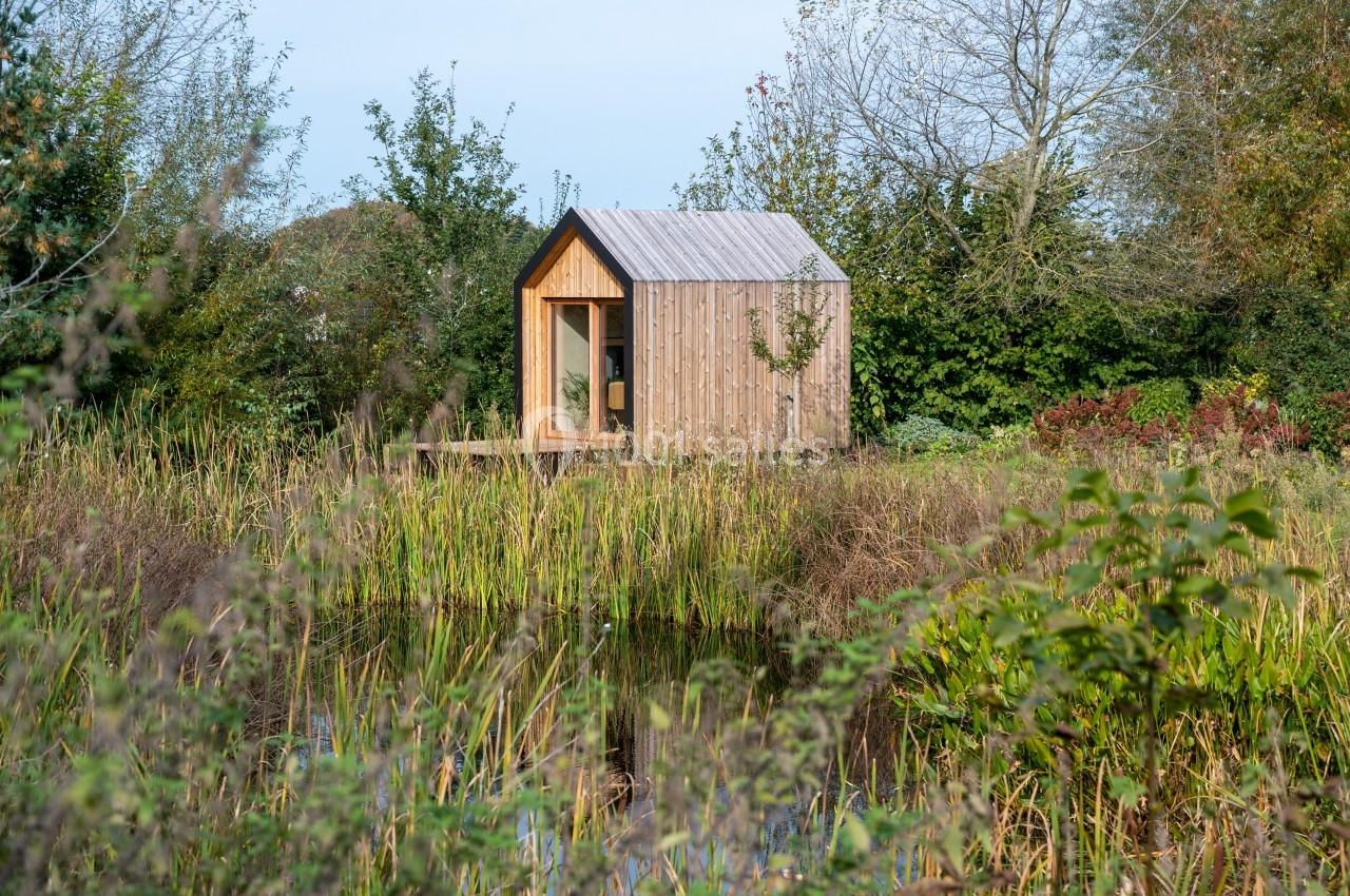 Cabane en bois au bord d'une mare, entourée de végétation et d'arbres sous un ciel clair.