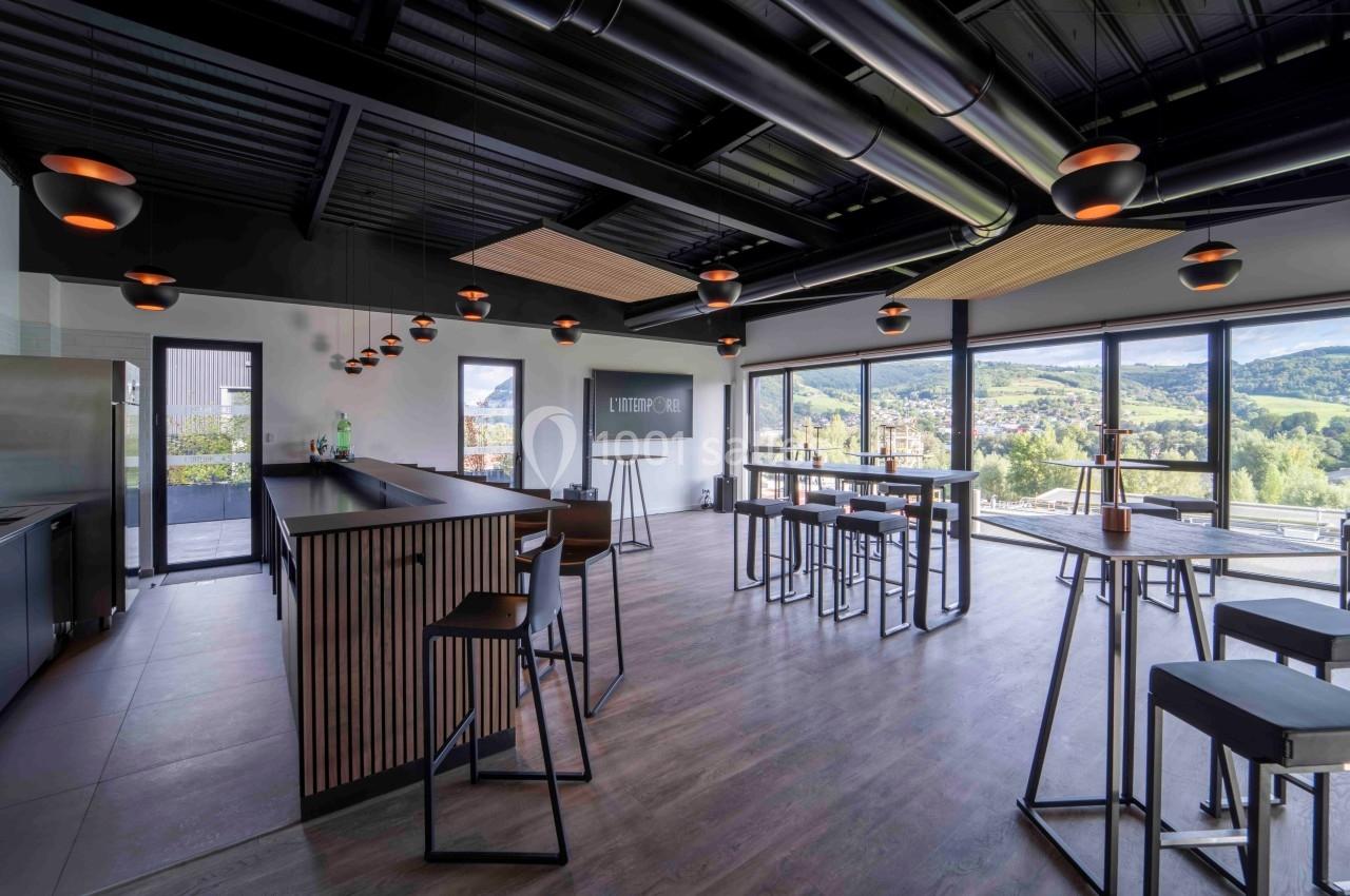 Salle moderne avec bar, tables hautes et grandes baies vitrées offrant une vue sur un paysage vallonné.
