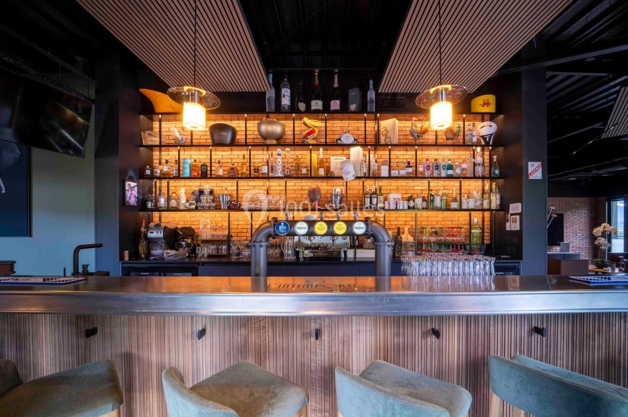 Bar moderne avec comptoir en bois, tabourets verts et étagères éclairées exposant des bouteilles et des verres.