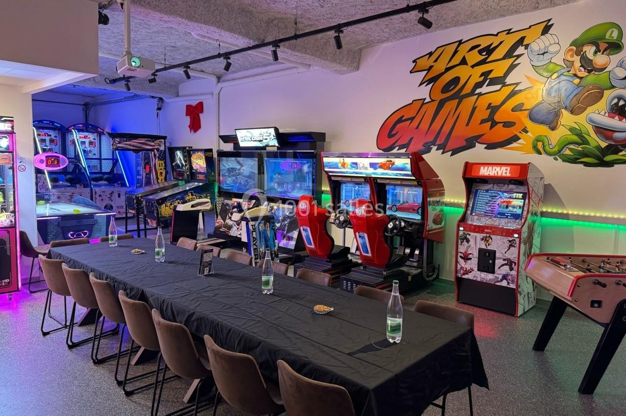 Salle d'arcade avec bornes de jeux, flippers, simulateurs de course et table dressée pour un événement.