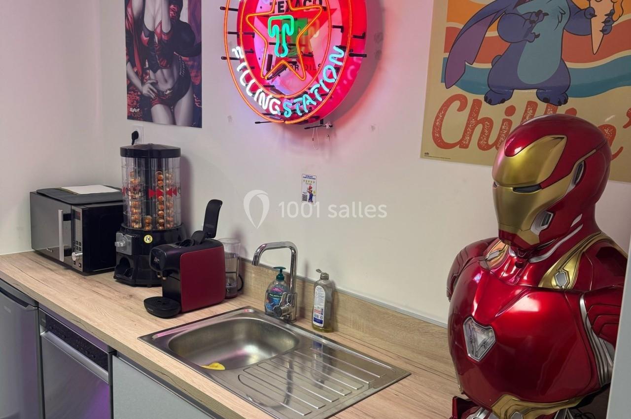 Cuisine équipée avec évier, cafetières, distributeur de bonbons, déco néon et posters, et buste d'armure rouge.