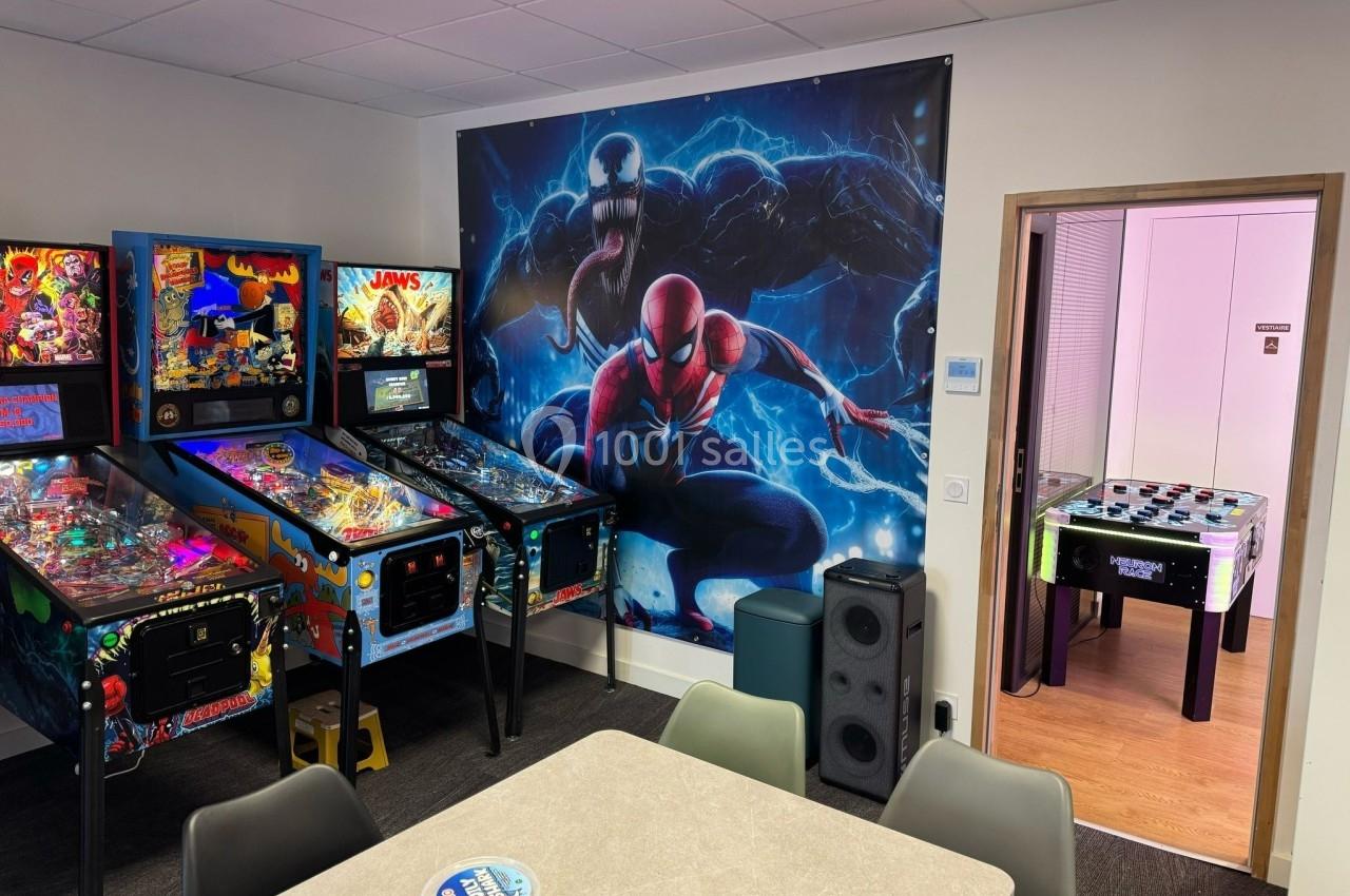 Salle de détente avec flippers, baby-foot, table et mur décoré d'une illustration de Spider-Man et Venom.