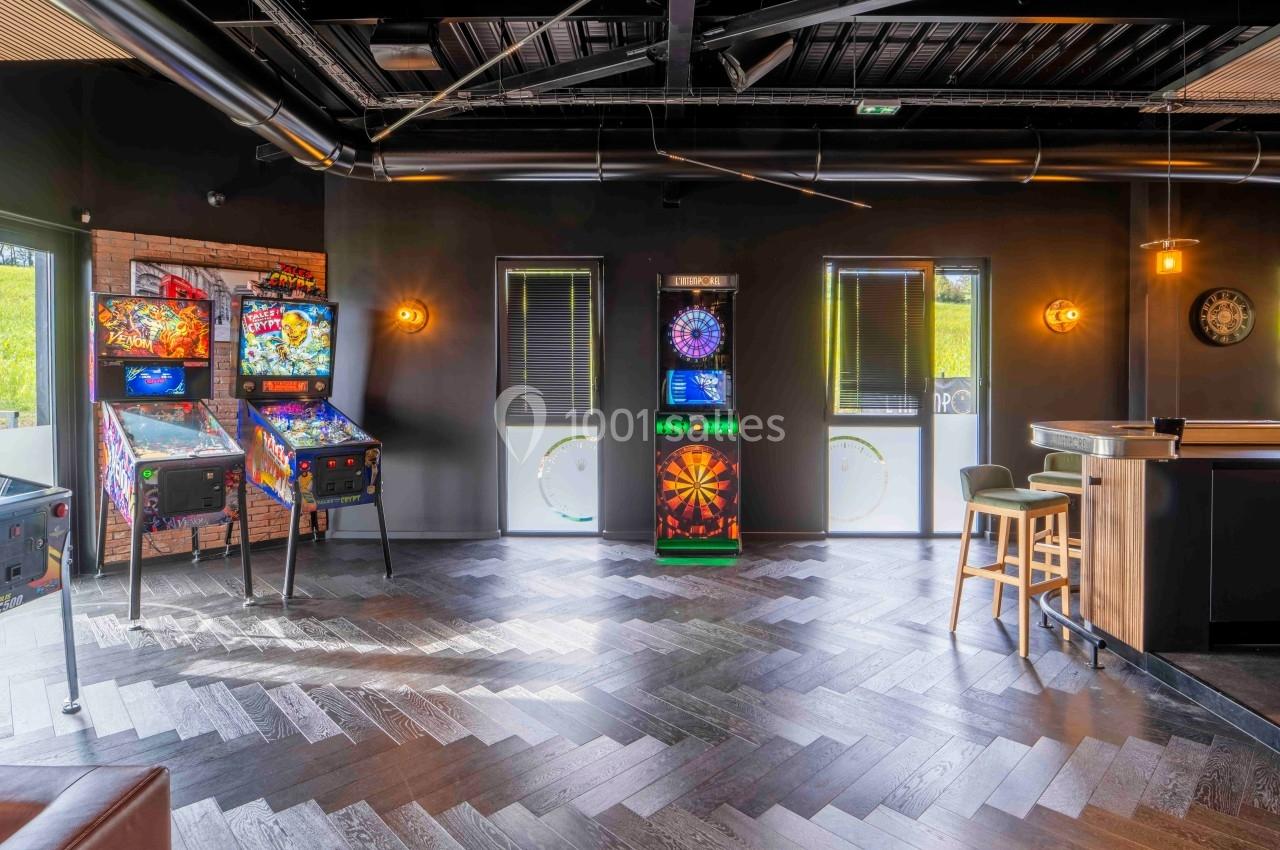 Salle de jeux avec flippers, cible de fléchettes électronique et bar dans un espace moderne et lumineux.