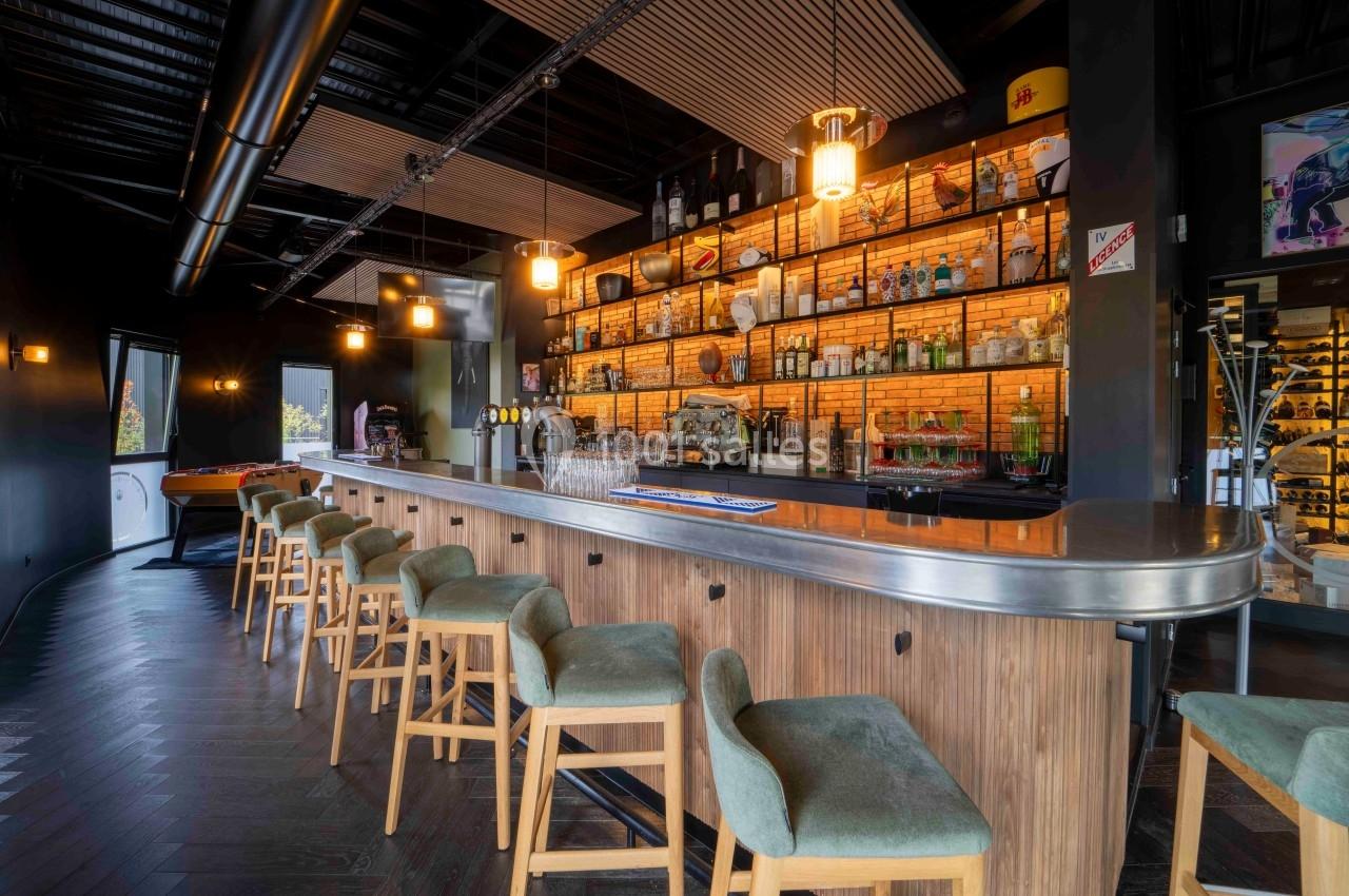 Bar moderne avec comptoir en bois, tabourets rembourrés, étagères éclairées contenant des bouteilles et ambiance chaleureuse.