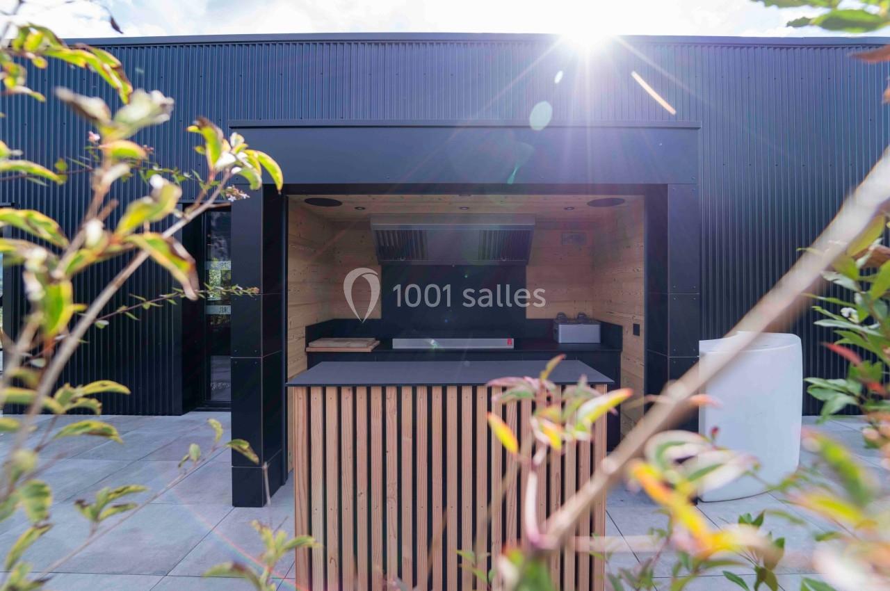 Cuisine extérieure moderne avec comptoir en bois, abri noir et lumière naturelle filtrée par le soleil.