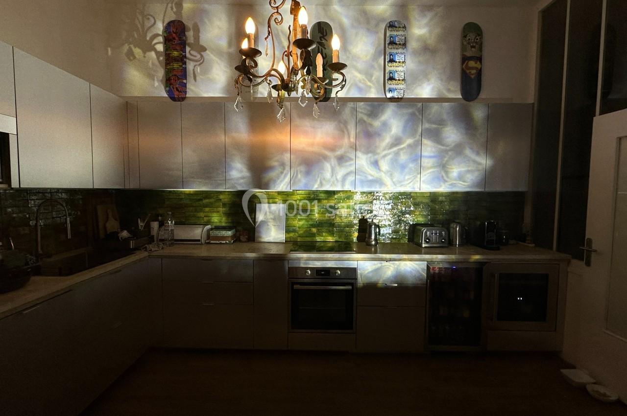 Cuisine moderne éclairée par un lustre, avec reflets lumineux sur les murs et des planches de skate accrochées en haut.