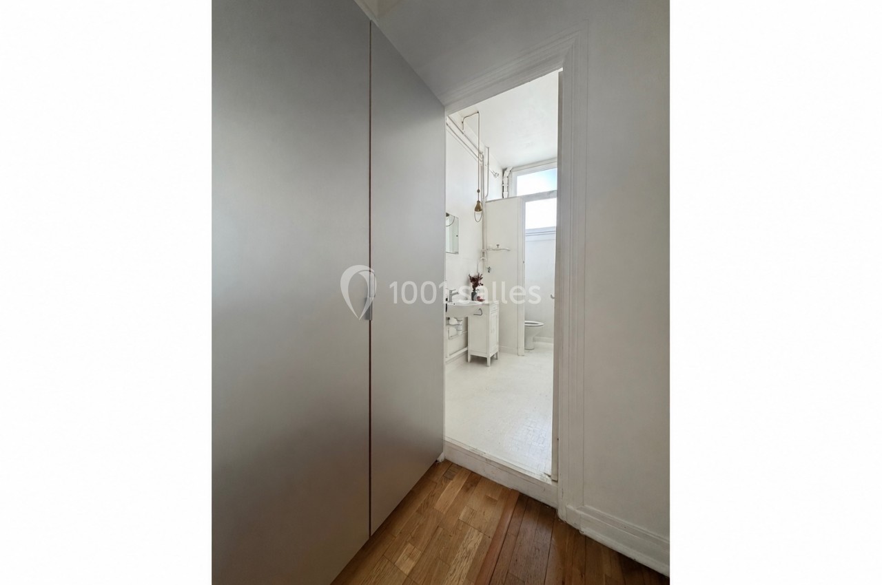 Vue d'une salle de bain blanche minimaliste avec lavabo, miroir et toilettes, prise depuis une pièce adjacente.