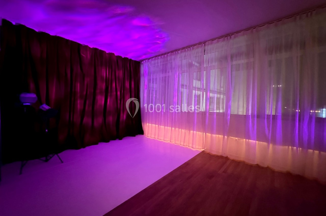 Salle avec rideaux violets et translucides éclairés par une lumière rose, sol en bois et mur blanc.