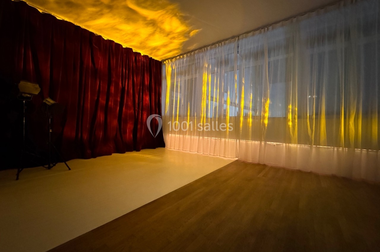 Salle avec rideaux rouges et blancs, éclairée par une lumière jaune, sol en bois et zone blanche au sol.