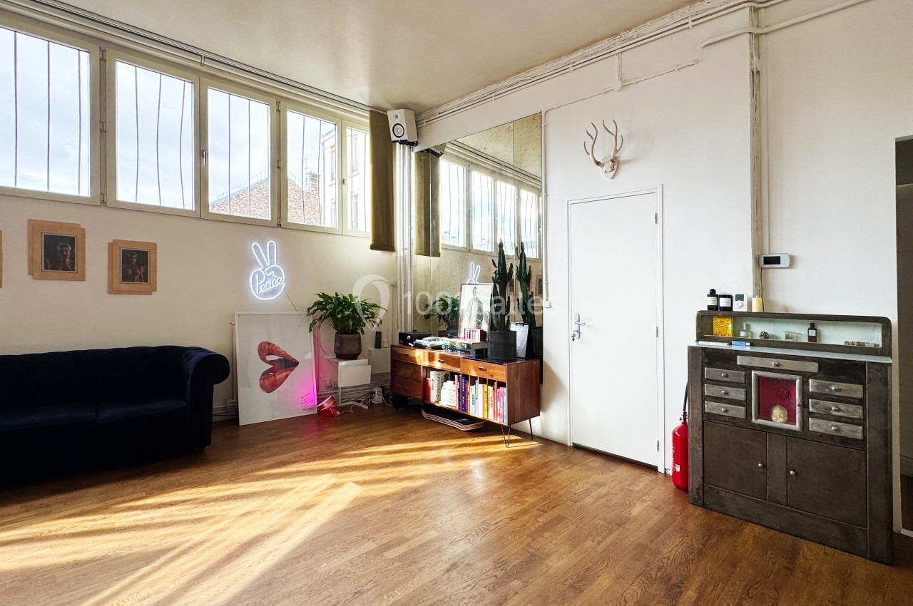 Salon lumineux avec parquet, canapé, meubles en bois, plantes, décorations murales et grande fenêtre vitrée.