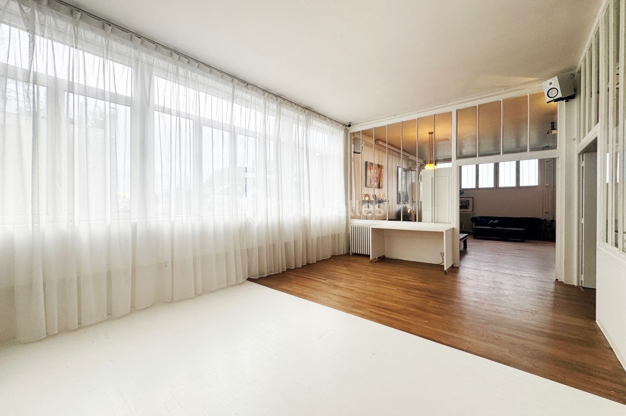 Salle lumineuse avec parquet et murs blancs, séparée par des verrières, éclairée par de grandes fenêtres avec rideaux.