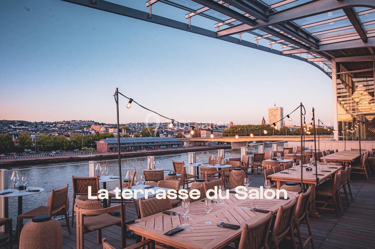 Terrasse aménagée avec tables et chaises en bois, vue sur une ville au bord d'un fleuve au coucher du soleil.