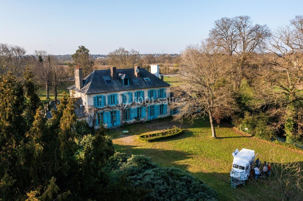 Manoir ancien avec volets bleus entouré d'un grand jardin arboré, un petit groupe près d'une camionnette blanche.