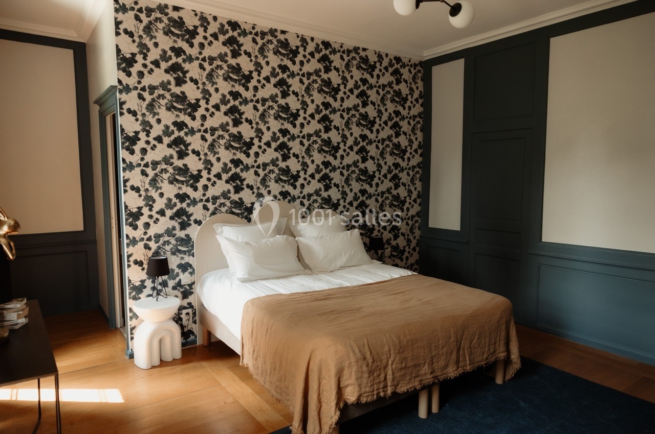 Chambre avec lit double, couvre-lit beige, mur tapissé de motifs floraux noirs et blancs, et parquet en bois clair.