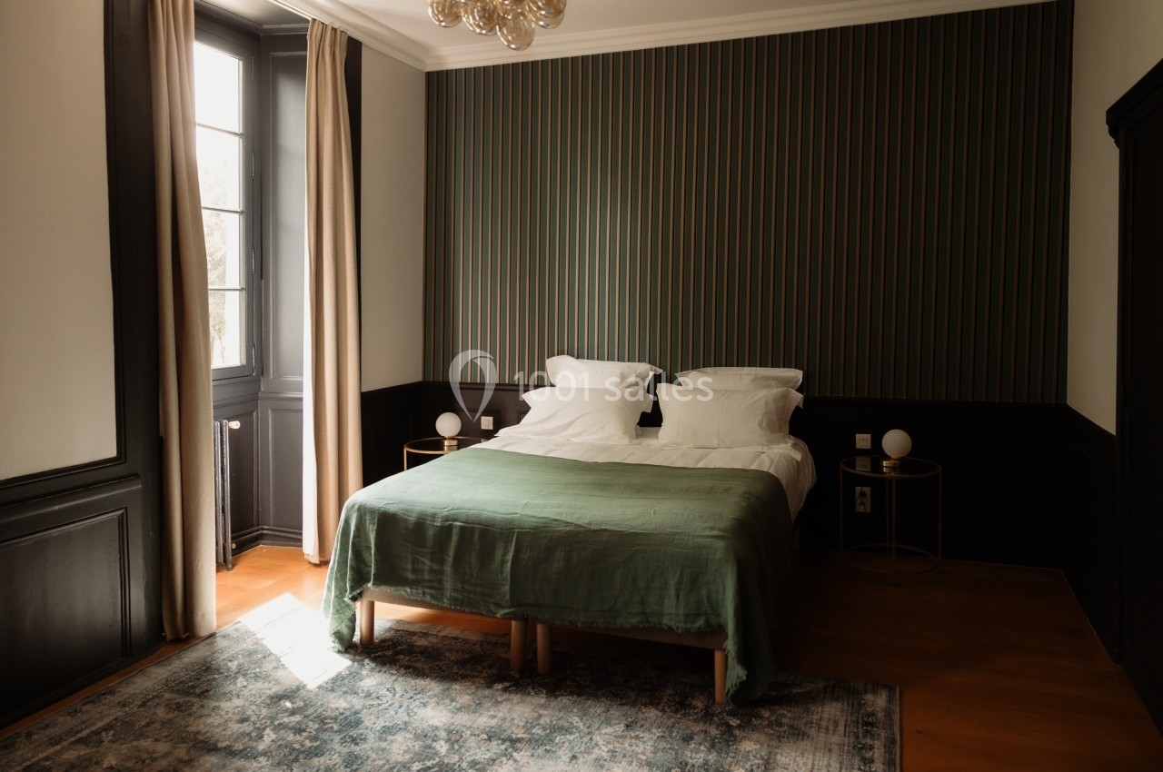 Chambre lumineuse avec lit double, couvre-lit vert, murs à rayures et grandes fenêtres avec rideaux beiges.