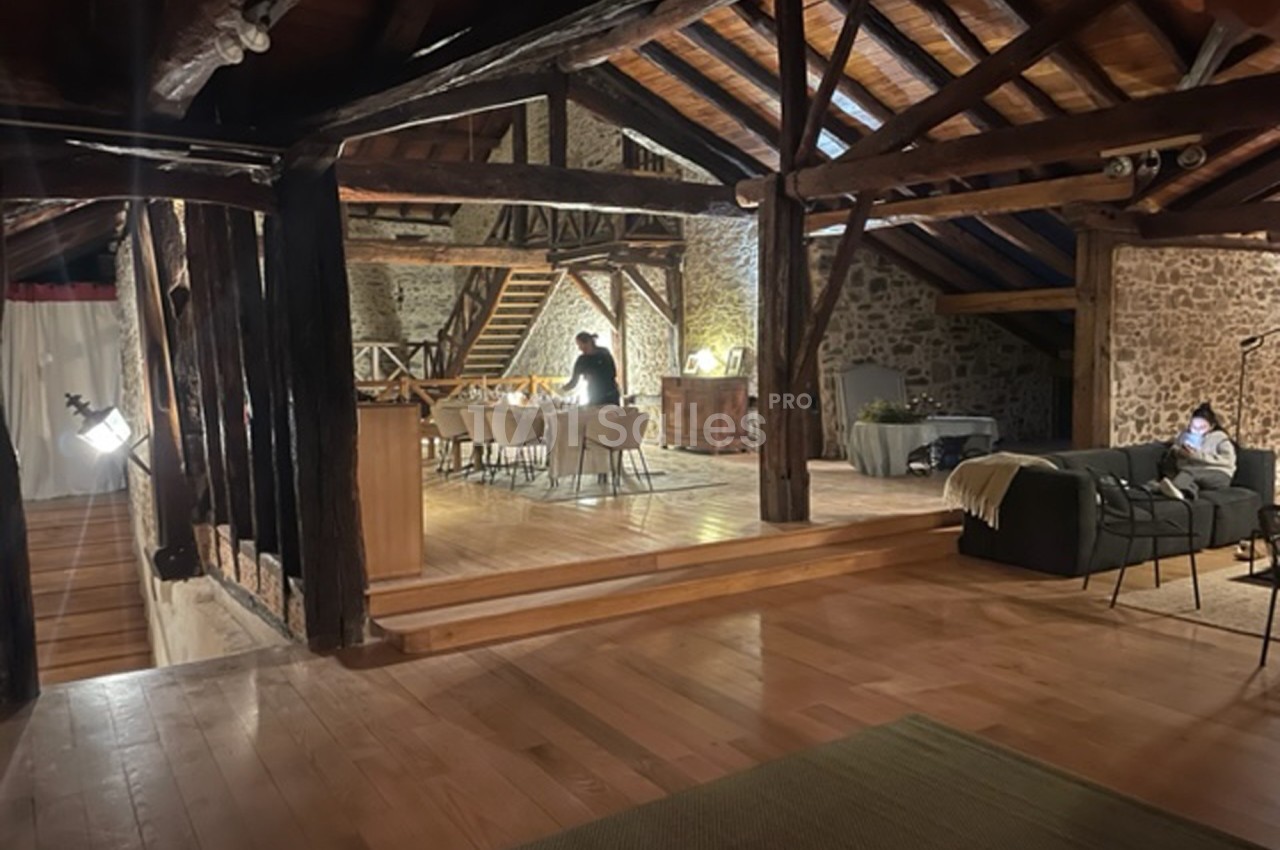 Vue d'un intérieur rustique avec poutres apparentes, espace repas éclairé et coin salon dans une ambiance chaleureuse.