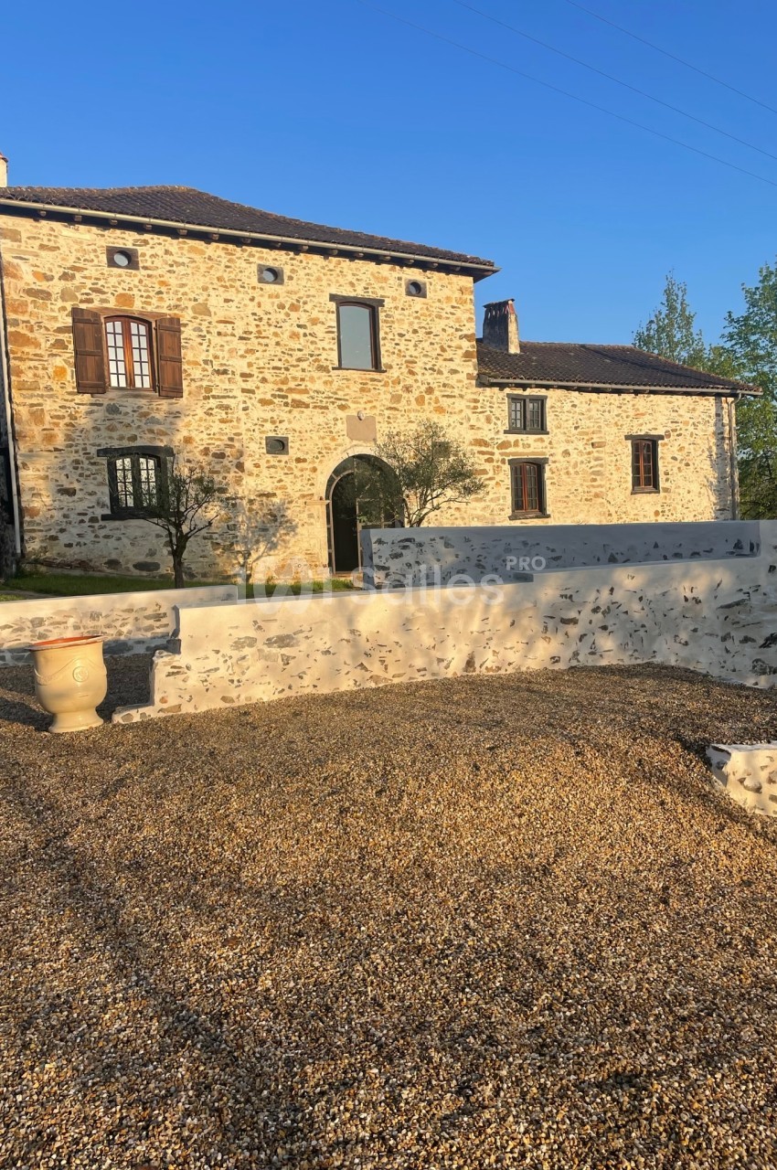 Maison en pierre avec volets en bois, entourée d'une cour gravillonnée et d'un mur bas, sous un ciel dégagé.