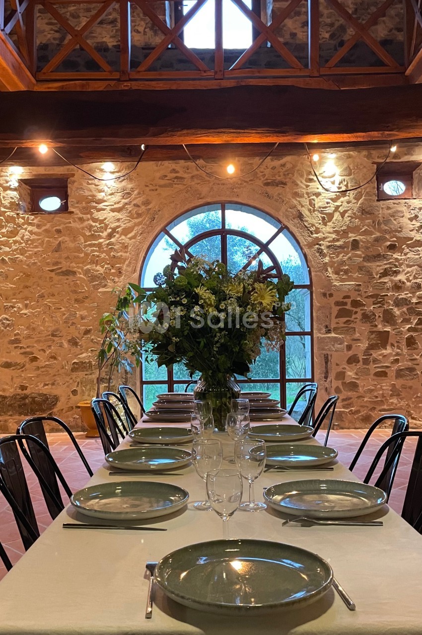 Grande table dressée avec vaisselle et bouquet de fleurs, dans une salle en pierre avec une grande fenêtre en arche.