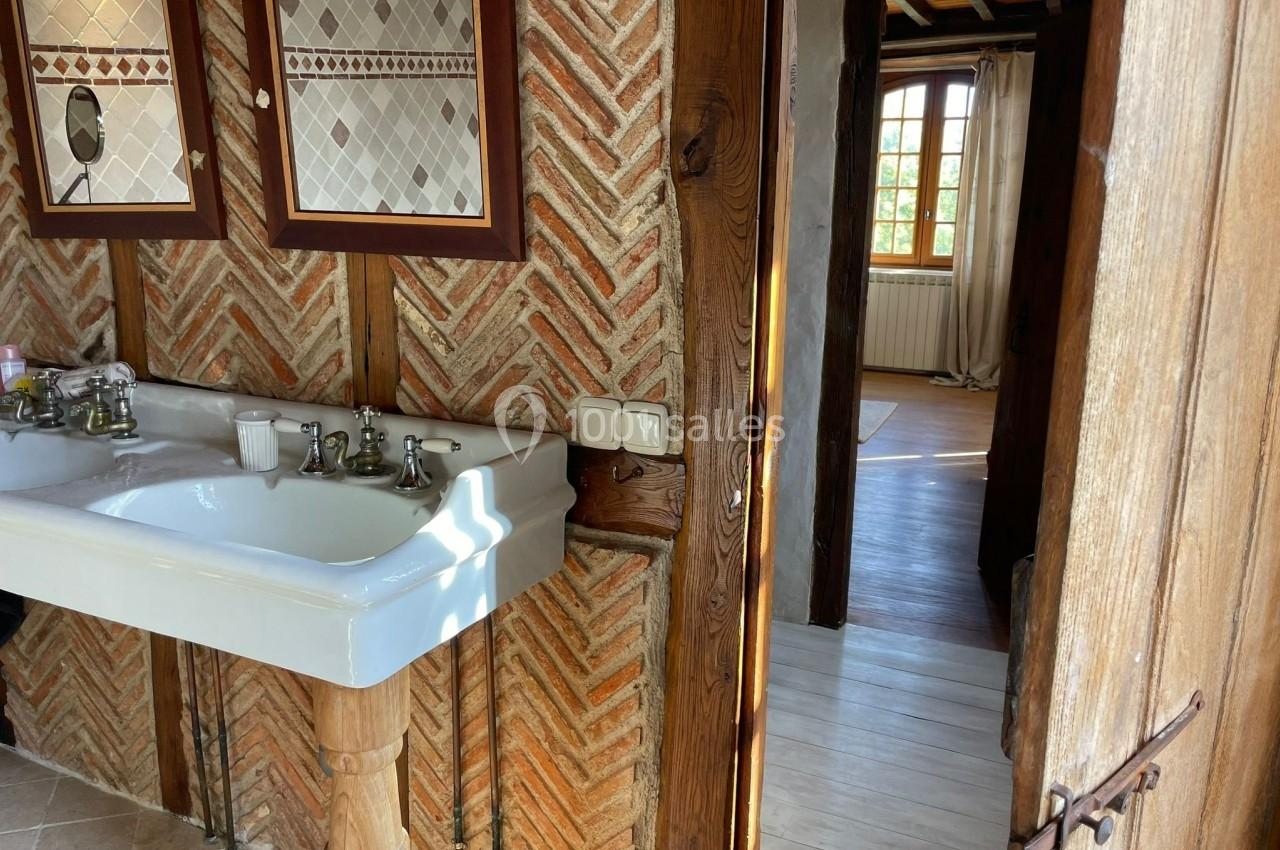 Double lavabo en céramique blanche avec robinets, installé contre un mur en briques, vue sur une pièce lumineuse adjacente.