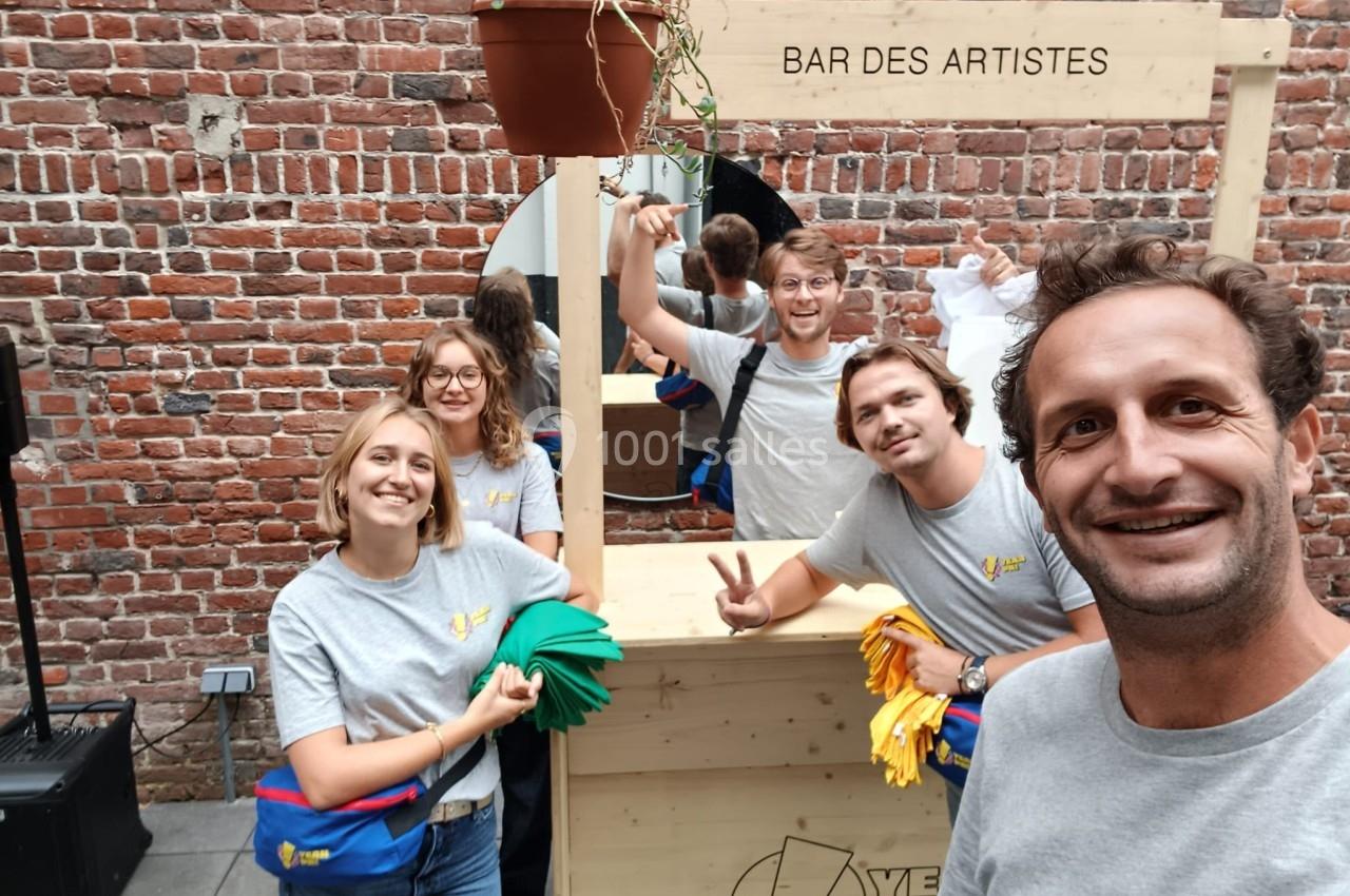 Des personnes souriantes posent devant un stand en bois marqué ’Bar des artistes’, dans une cour en briques.