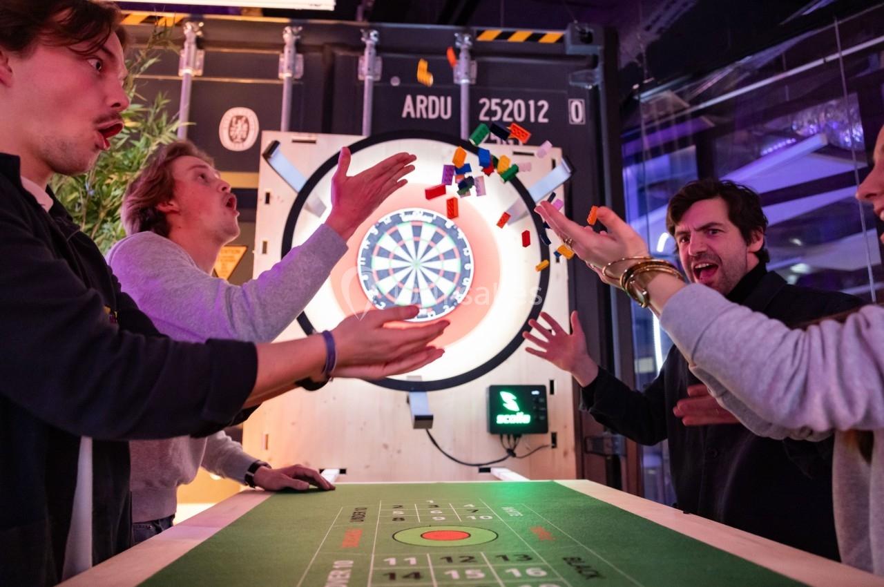 Des personnes jouent autour d'une table avec des jetons colorés, un jeu de fléchettes visible en arrière-plan.