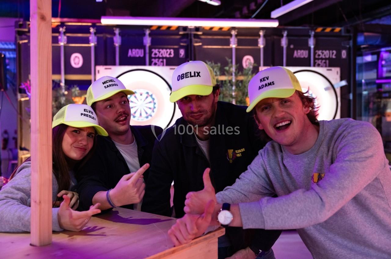 Quatre personnes souriantes portant des casquettes personnalisées posent devant un jeu de fléchettes lumineux.