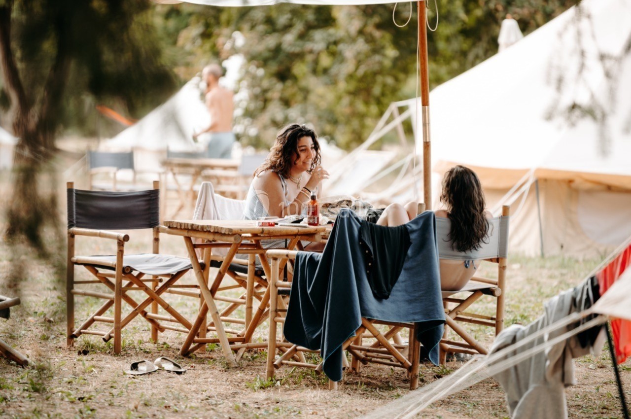 Deux femmes discutent à une table en bois sous un parasol, dans un espace extérieur avec des tentes en arrière-plan.