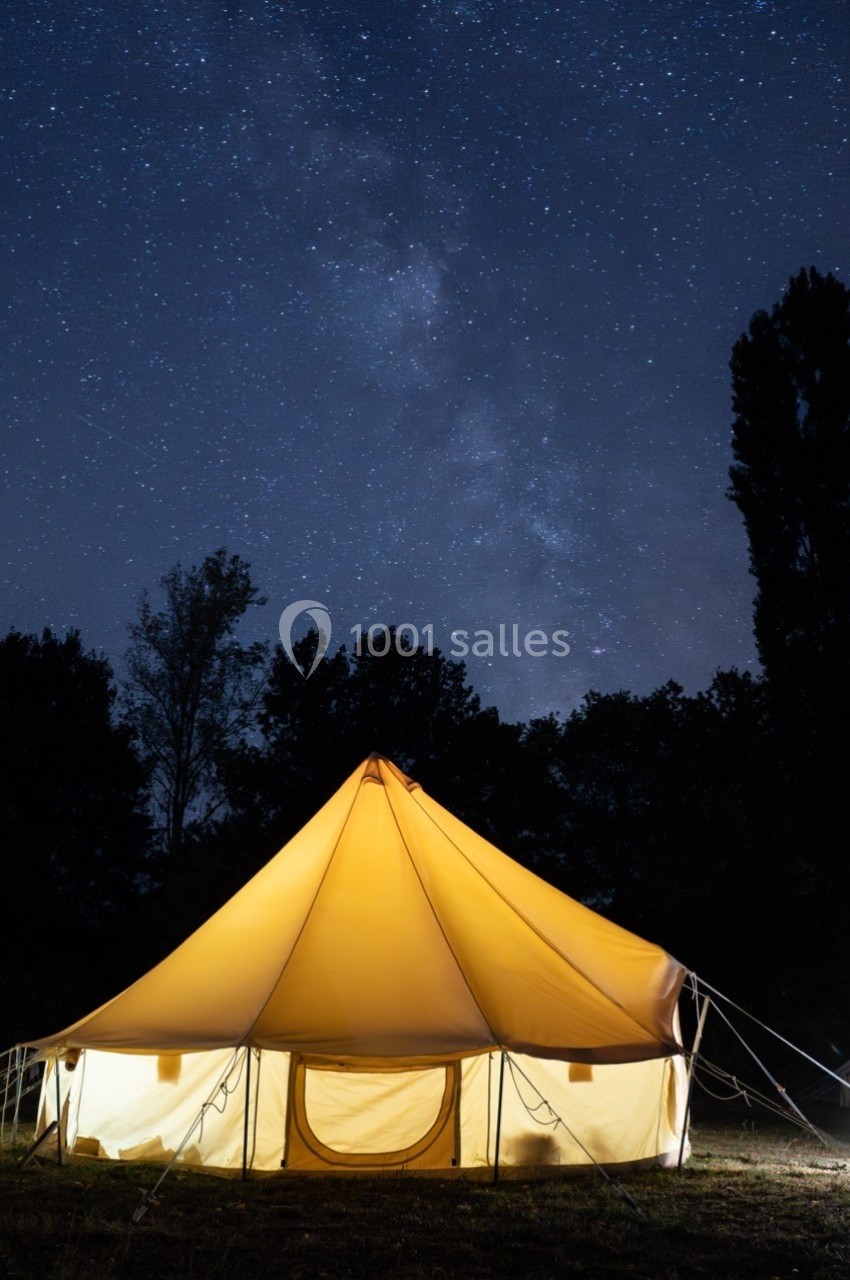 Tente éclairée sous un ciel étoilé, entourée d'arbres dans un environnement nocturne paisible.