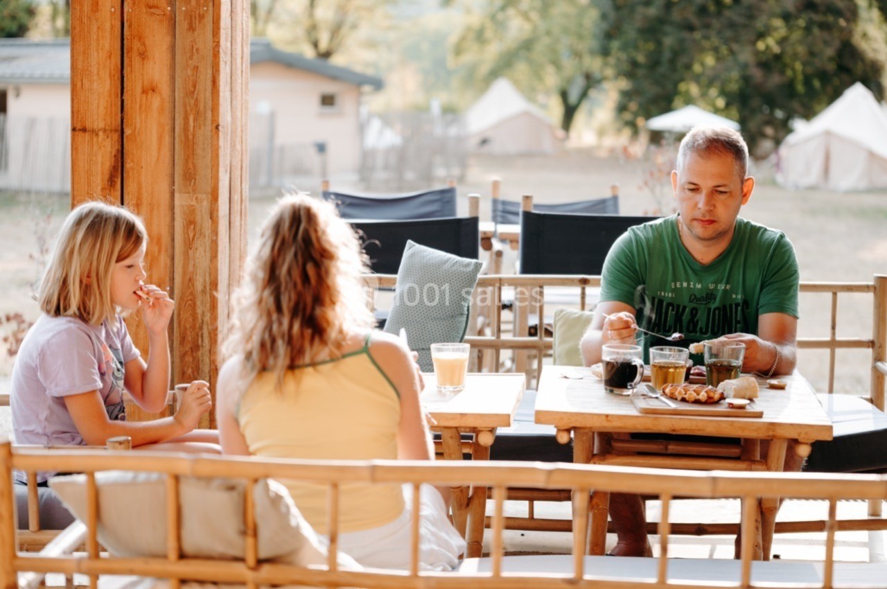 Une famille prend un petit-déjeuner en terrasse, entourée de mobilier en bois et d'un paysage de camping.