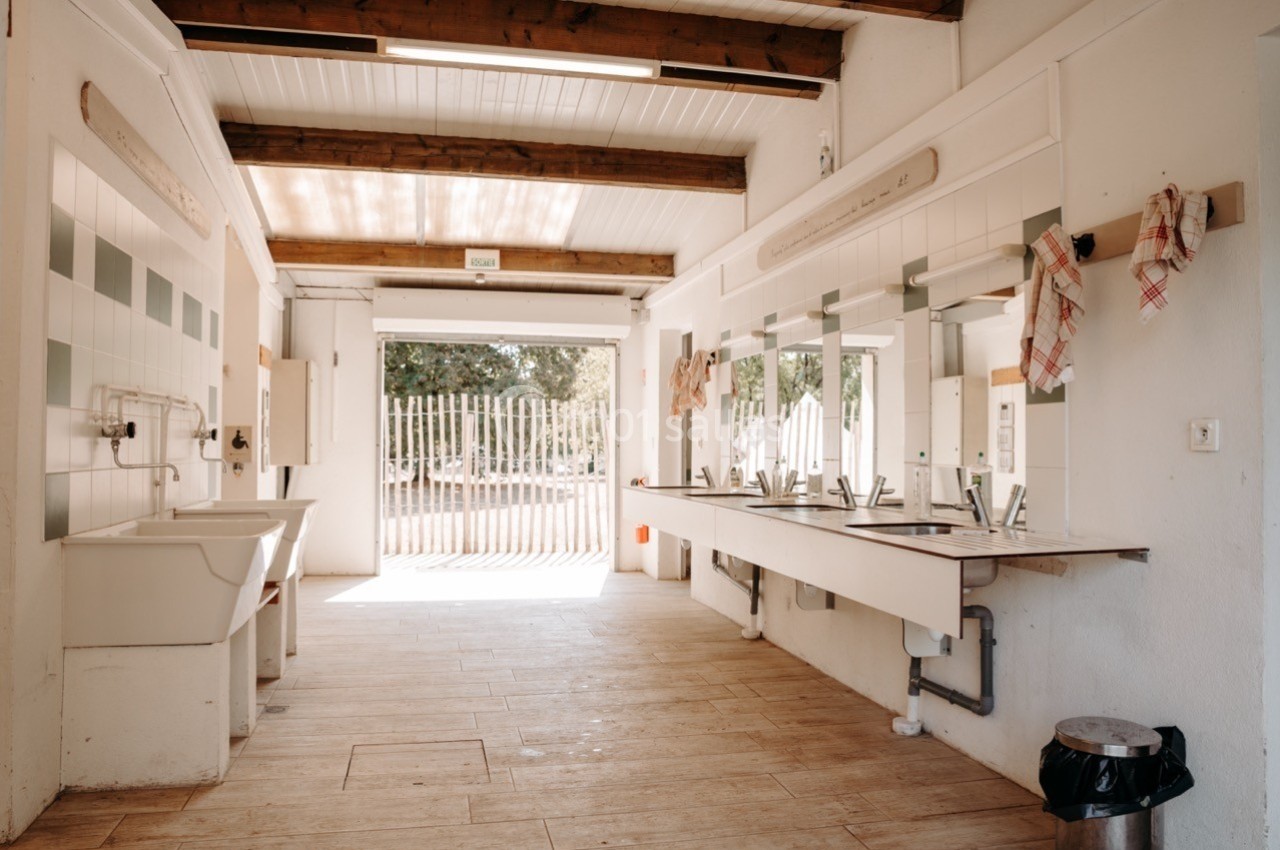 Intérieur d'un bloc sanitaire lumineux avec éviers, miroirs, lavabos et linge suspendu, ouvert sur l'extérieur.