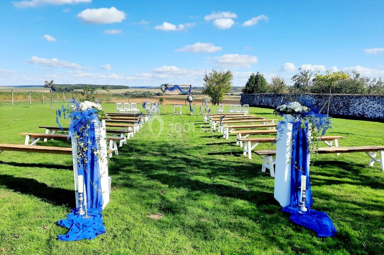 Allée centrale décorée de fleurs et tissus bleus menant à une arche pour une cérémonie en plein air sur une pelouse.