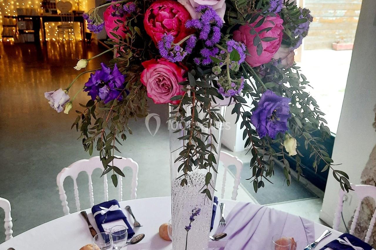 Centre de table fleuri avec roses, pivoines et feuillage, posé sur un vase haut, dans une salle décorée pour un événement.