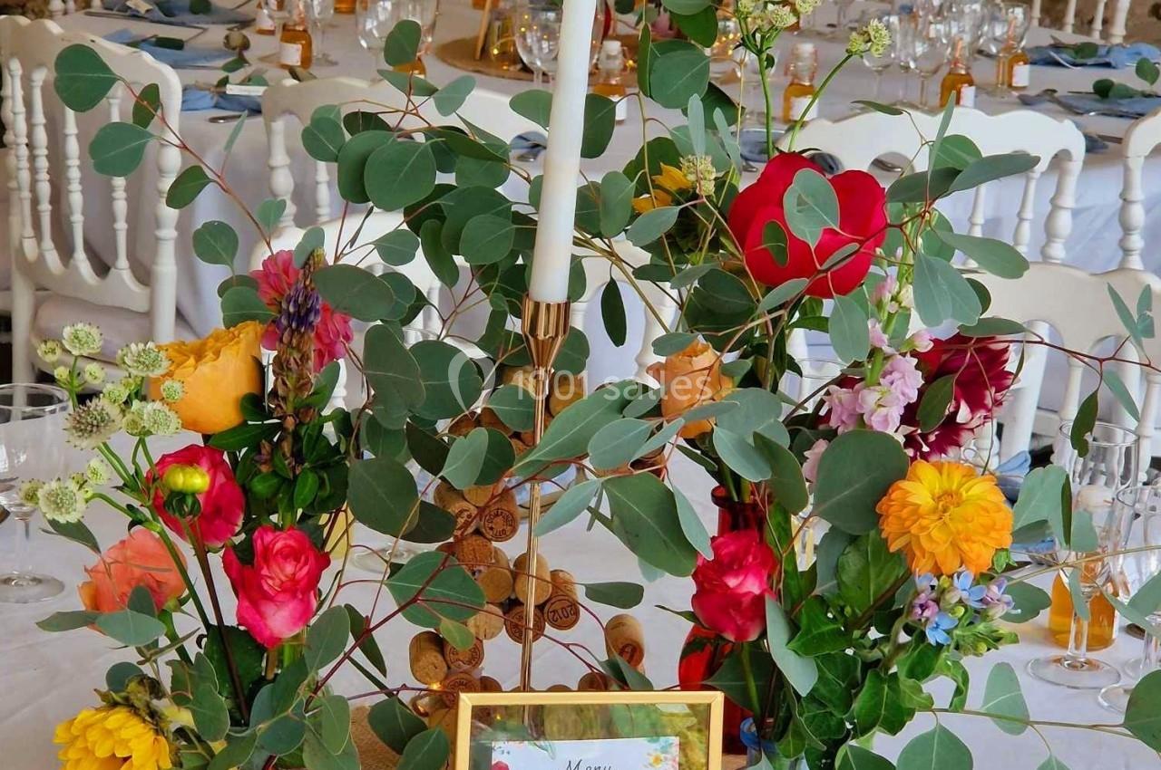 Centre de table floral coloré avec bougie blanche, menu encadré et décor rustique en arrière-plan.