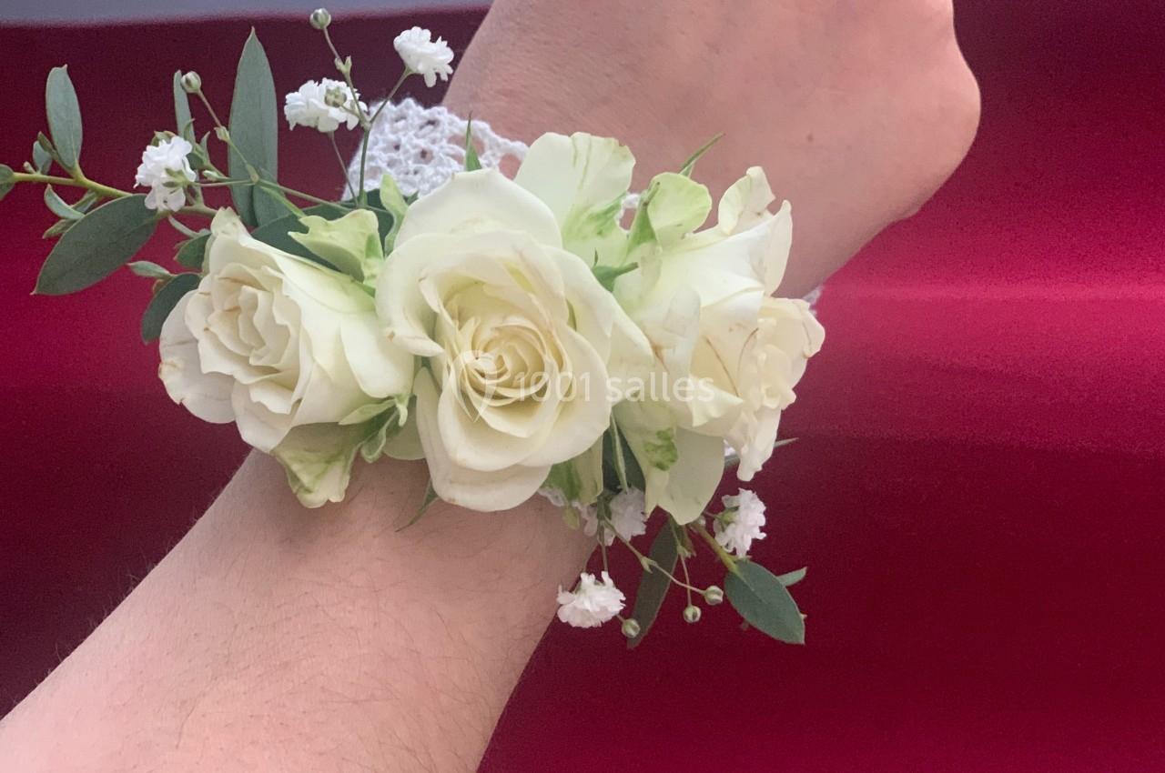 Bracelet floral composé de roses blanches et de gypsophiles, porté sur un poignet devant un fond rouge.