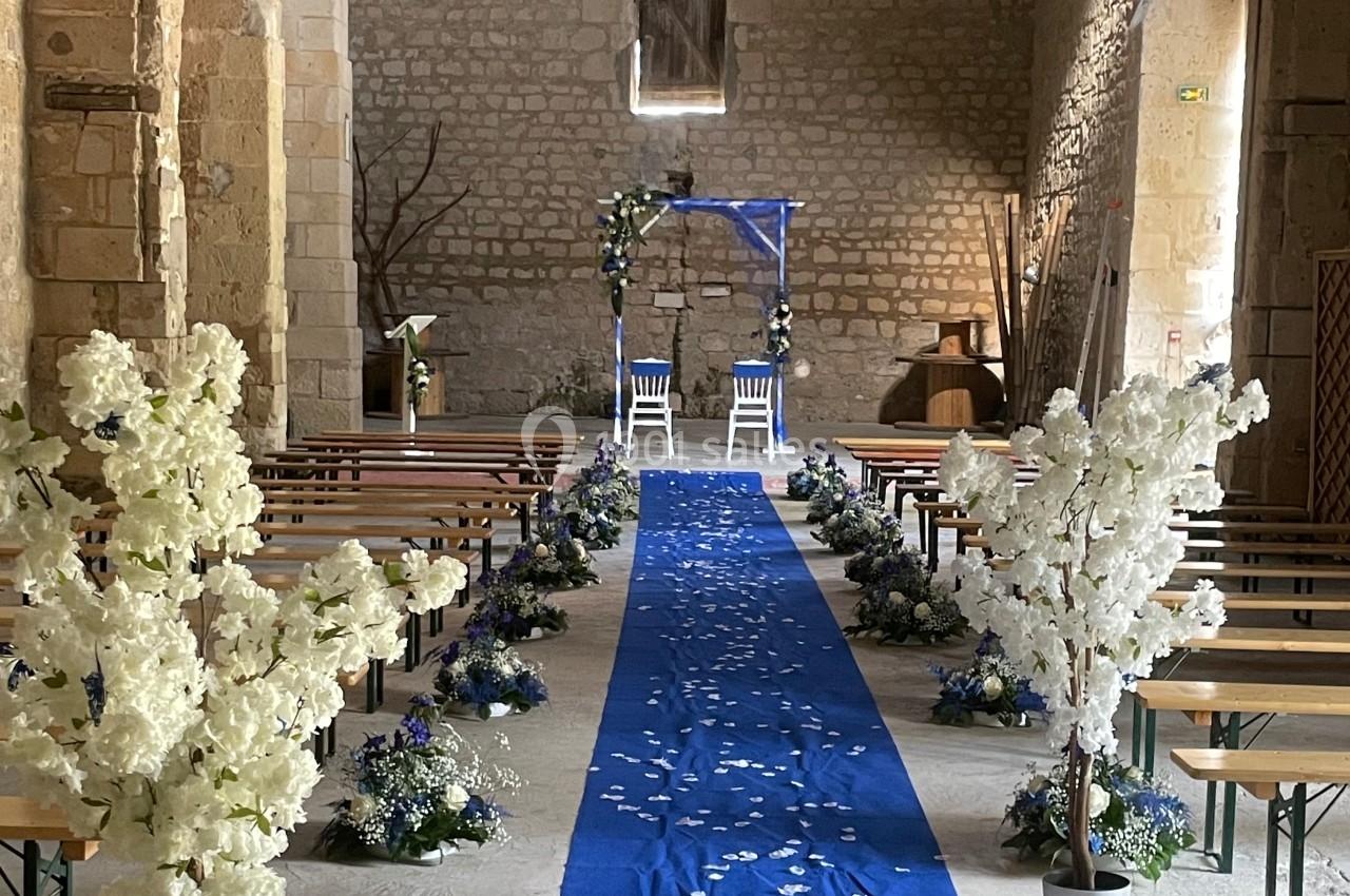 Allée centrale décorée de pétales blancs et tapis bleu menant à une arche fleurie dans une salle en pierre.