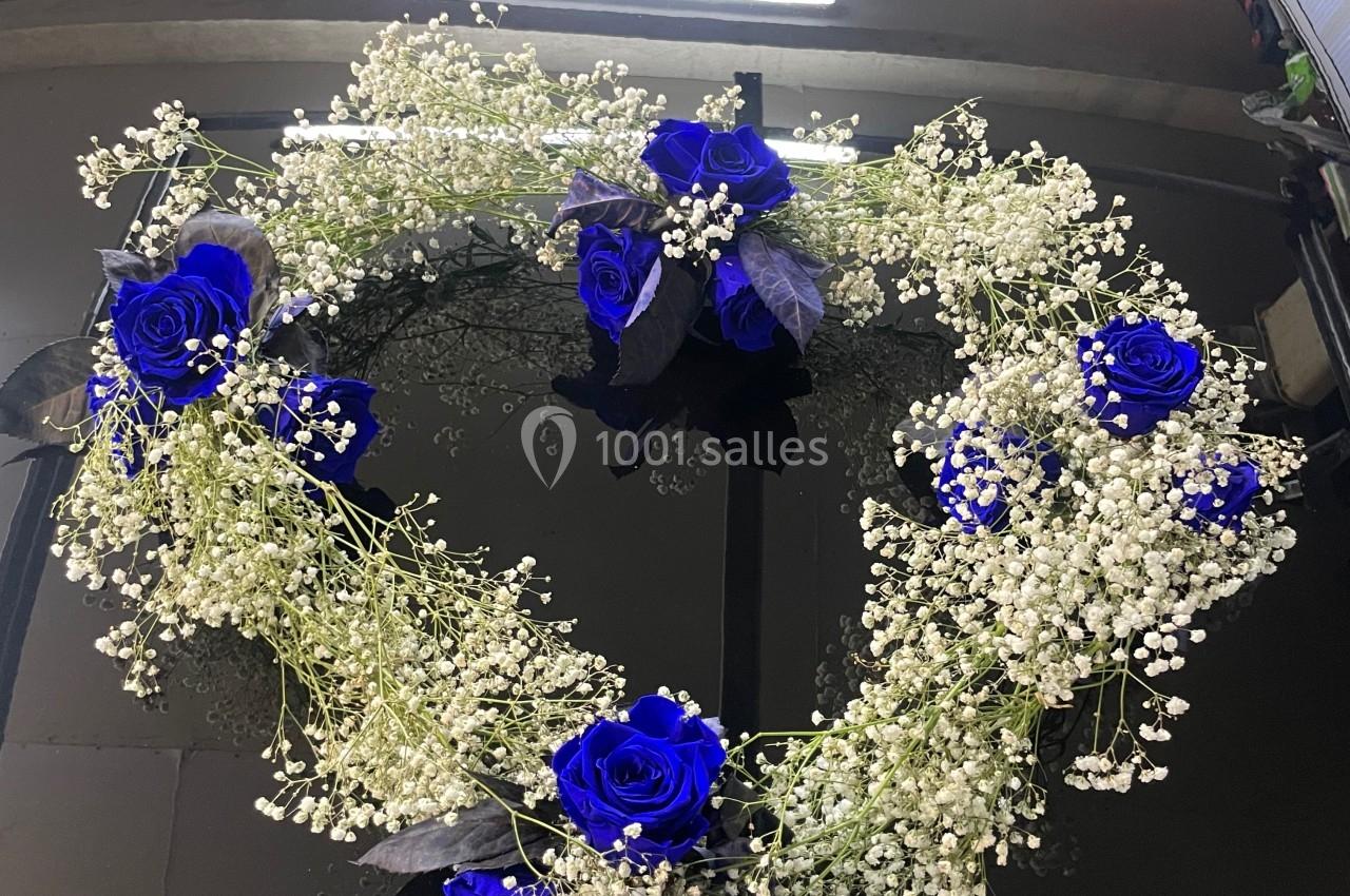 Cœur floral composé de roses bleues et de gypsophiles blanches posé sur le capot d'une voiture noire.