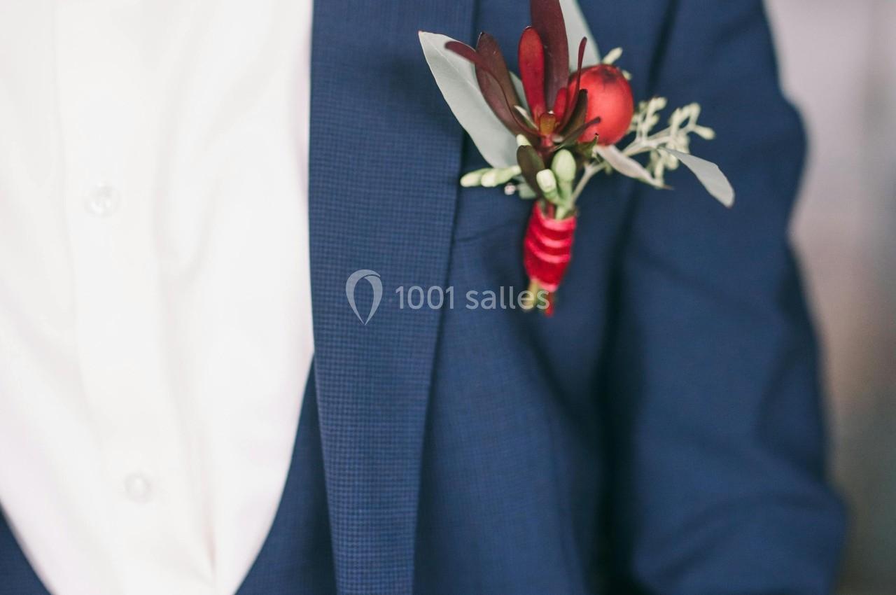 Homme en costume bleu avec une boutonnière rouge et blanche, portant un nœud papillon assorti.