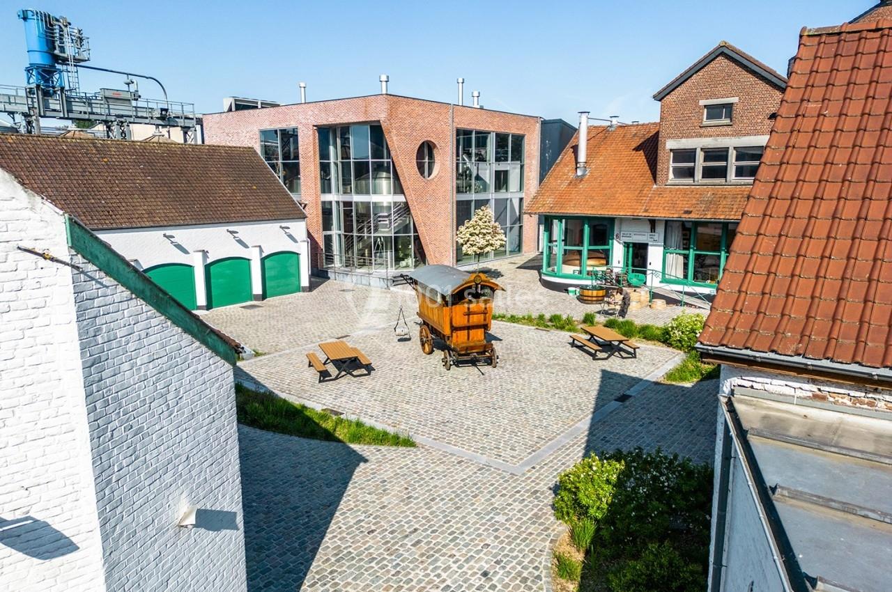 Brasserie Dubuisson Cour pavée entourée de bâtiments en briques et en crépi, avec une roulotte en bois et des bancs au centre.