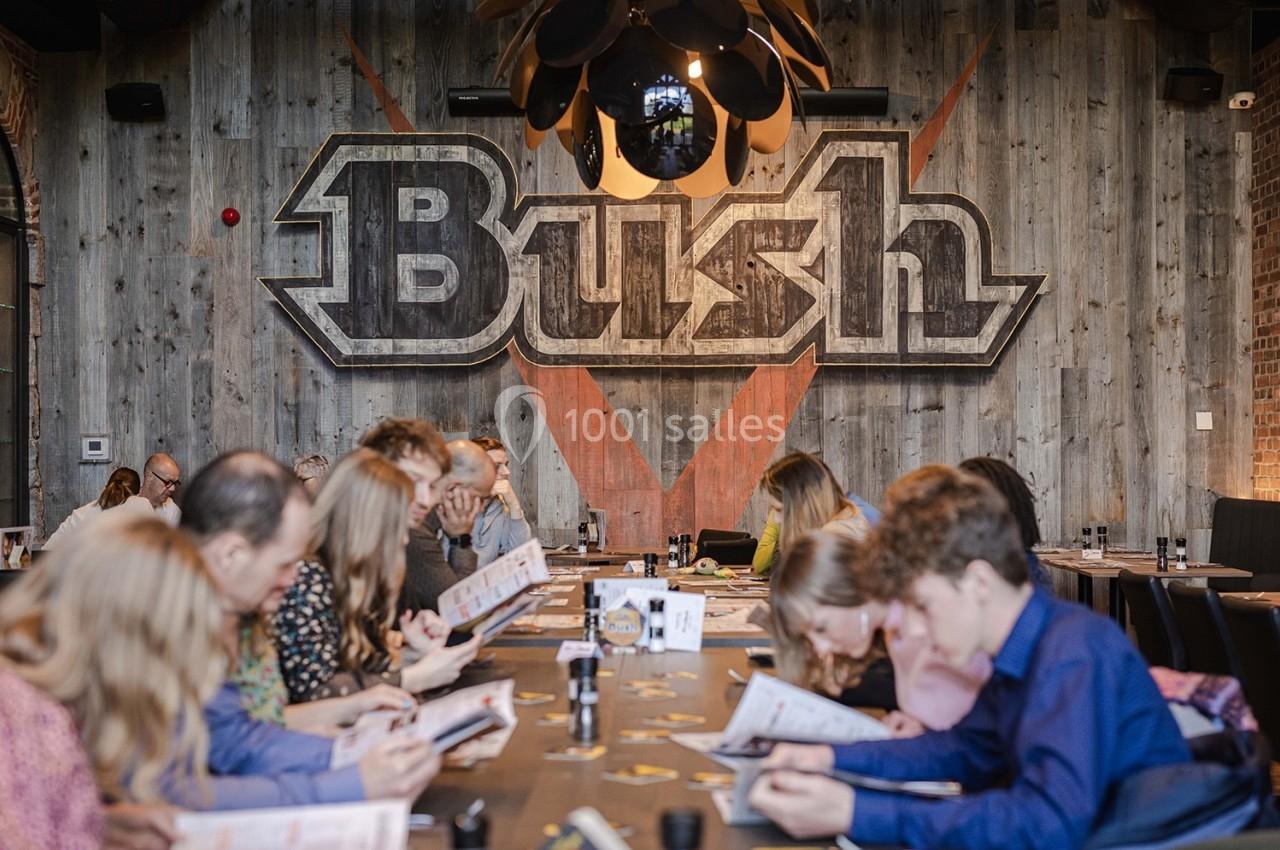 Brasserie-taverne Trolls&Bush Des personnes assises autour d'une grande table dans un restaurant, lisant des menus devant un mur décoré du logo ’Bush’.