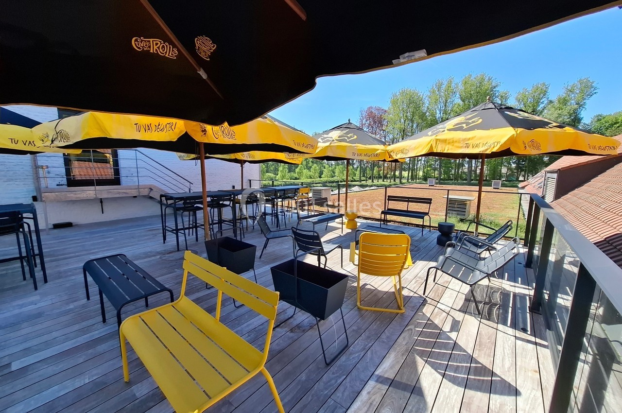 Rooftop Lowa Trolls Terrasse en bois avec tables, bancs jaunes et parasols jaunes et noirs, entourée de verdure par une journée ensoleillée.