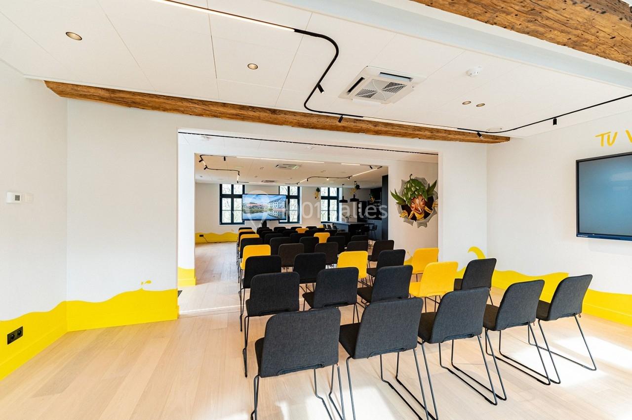 Salle Trolls Salle de réunion lumineuse avec rangées de chaises noires, écran mural et poutres apparentes.