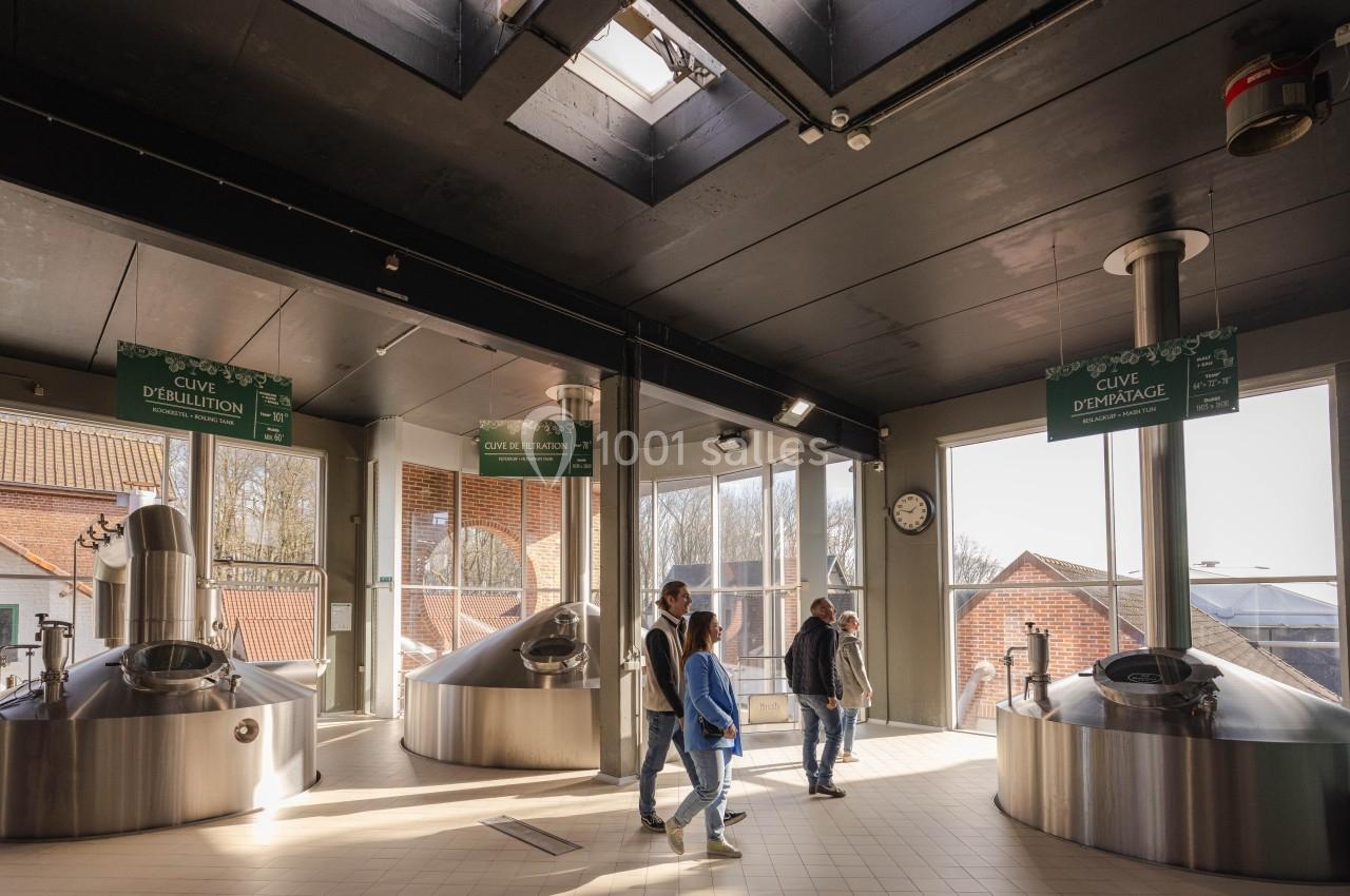 Visite de la Brasserie Visiteurs marchant dans une brasserie lumineuse avec cuves en acier inoxydable et panneaux explicatifs suspendus.