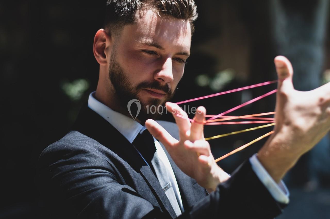 Un homme en costume joue avec des fils colorés tendus entre ses doigts dans un environnement extérieur ombragé.