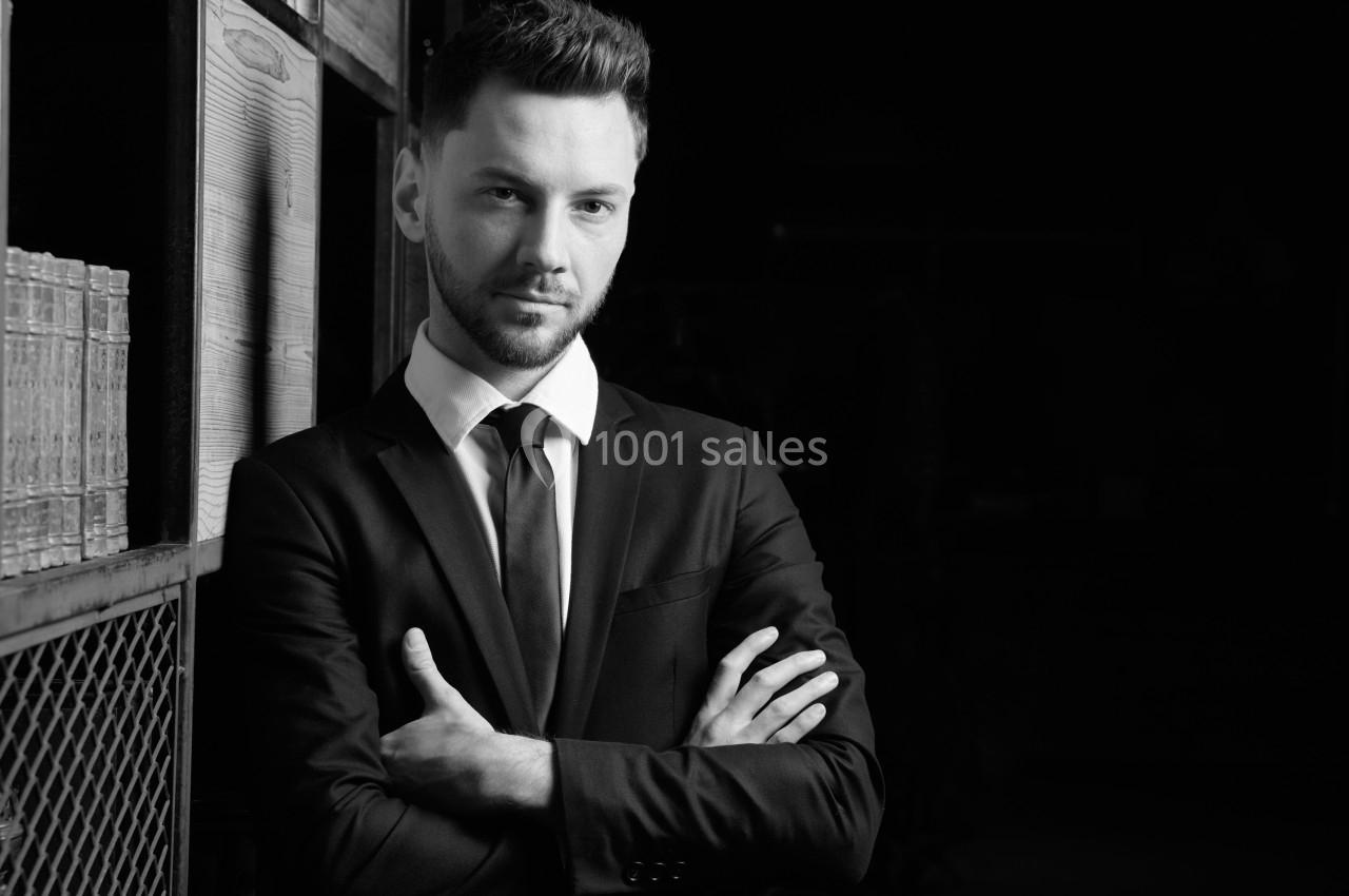 Homme en costume croisant les bras, debout près d'une étagère dans un environnement sombre.