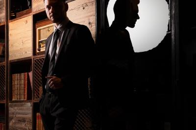 Homme en costume debout près d'une bibliothèque en bois, avec son ombre projetée sur un mur éclairé.