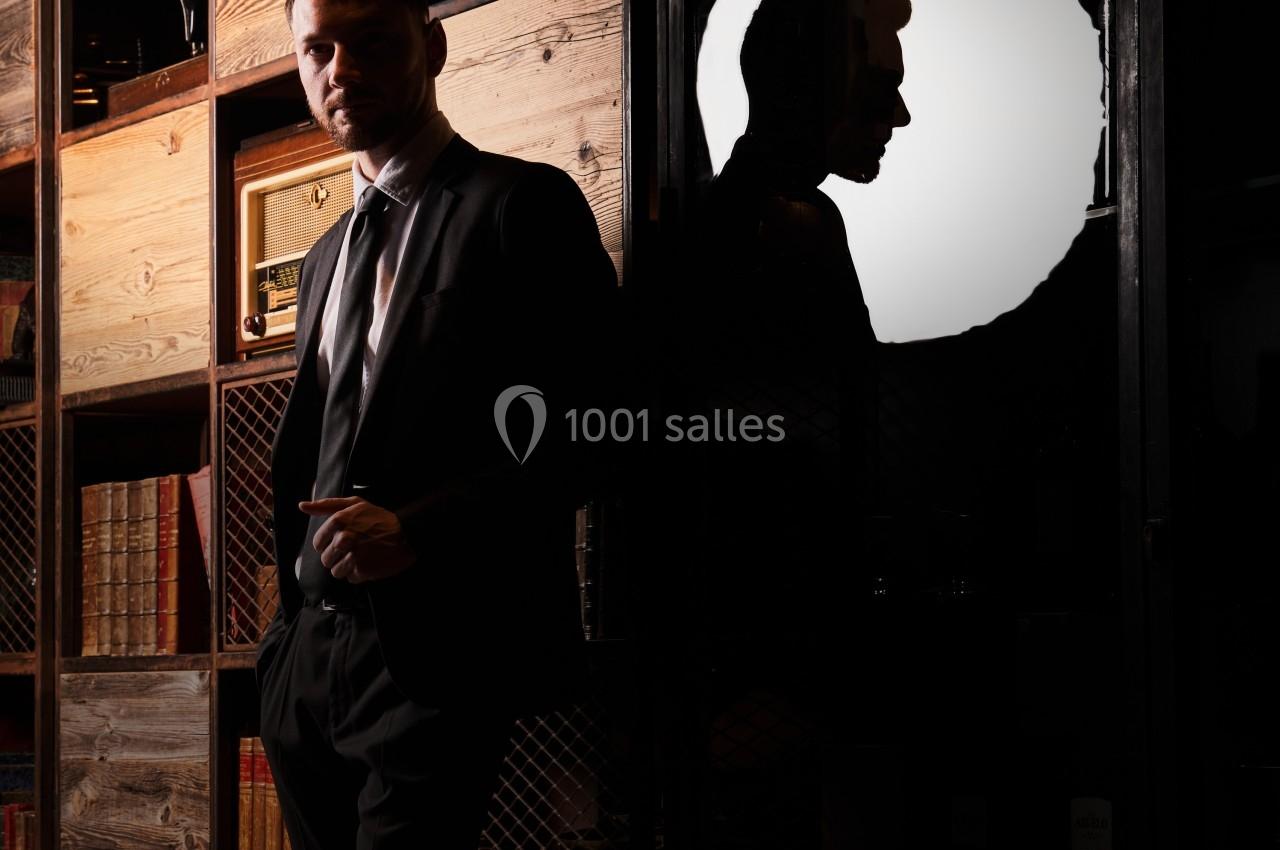 Homme en costume debout près d'une bibliothèque en bois, avec son ombre projetée sur un mur éclairé.