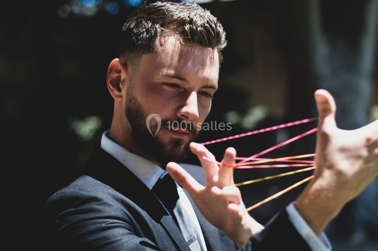 Un homme en costume manipule des fils colorés tendus entre ses doigts dans un environnement extérieur.